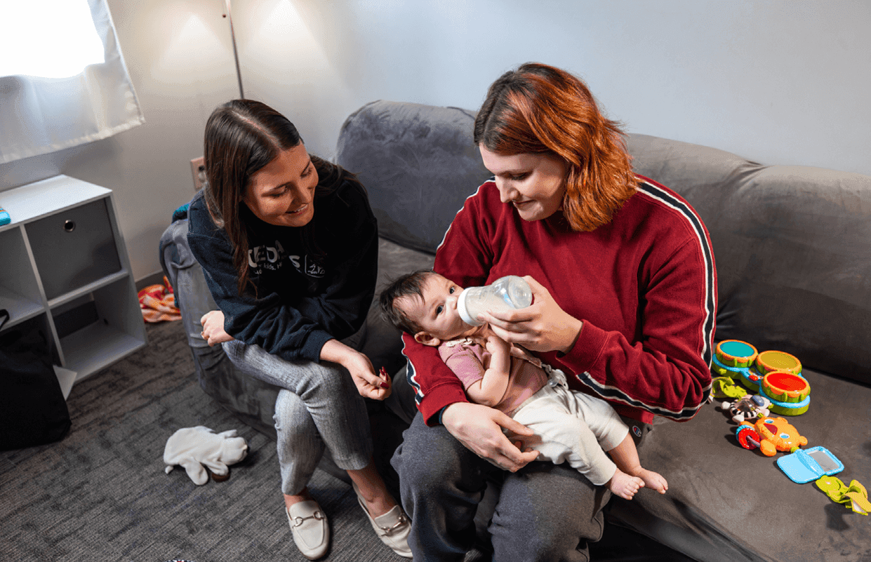 Young mom feeding her baby a bottle while a CEDARS staff member helps