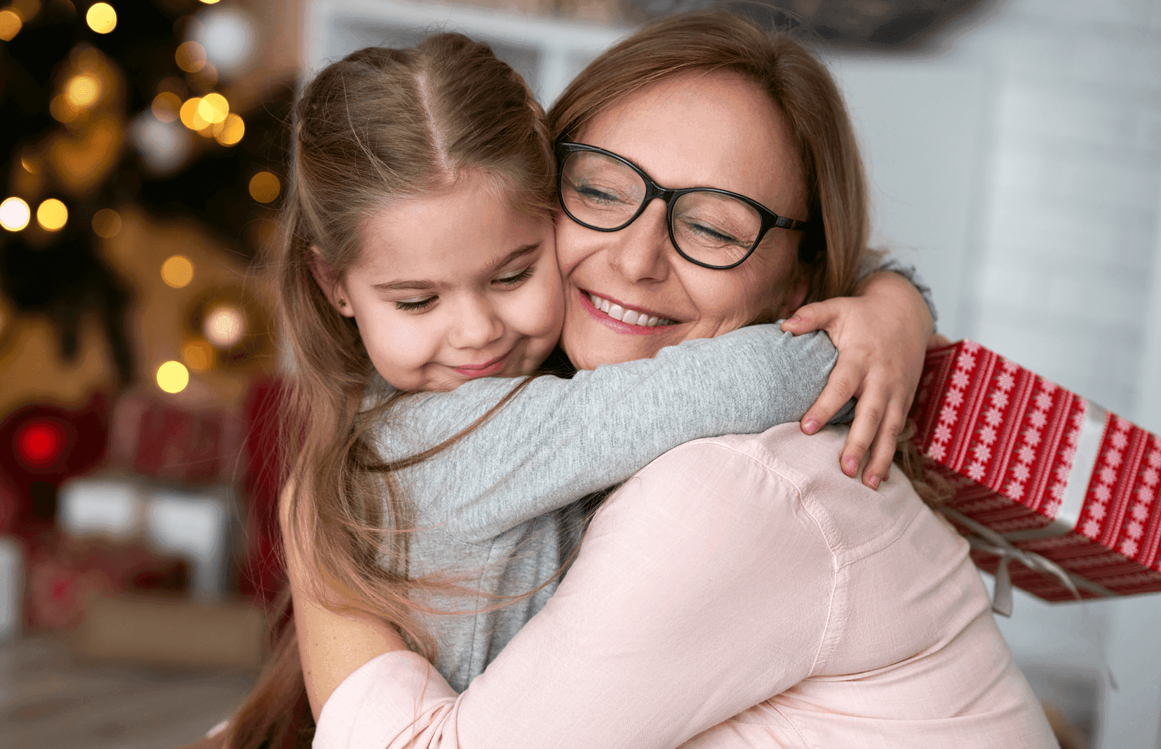 Middle aged woman with glasses hugging a young girl who is holding a holiday gift.