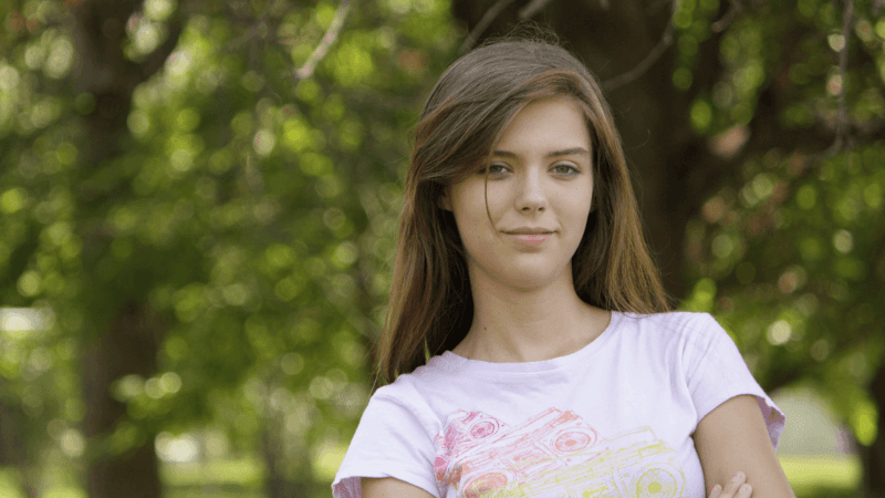 Teen girl with long brown hair and a pink shirt standing outside with her arms crossed while softly smiling at the camera